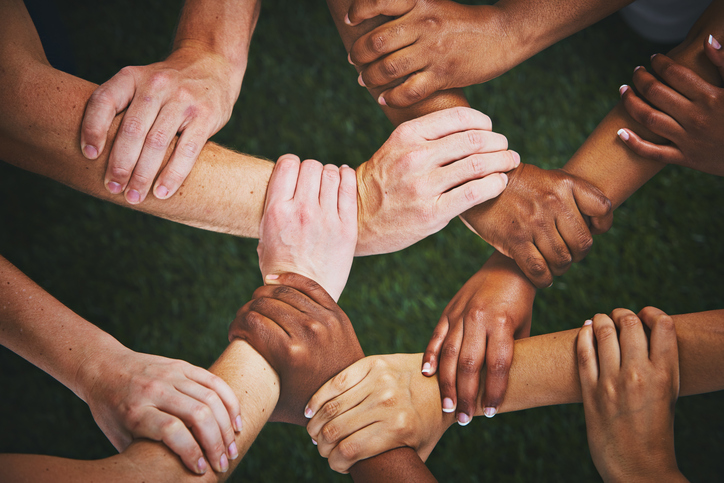 Looking down at many mixed hands, each clasping the wrist of another person and forming an interlinked human chain or mesh. Diversity
